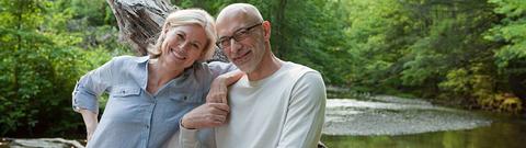 Mann und Frau schauen lächelnd in die Kamera, im Hintergrund Wald und Bach